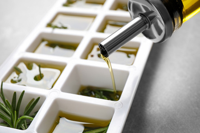 Photo of Pouring olive oil into ice cube tray with fresh rosemary at grey table, closeup