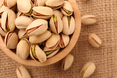 Photo of Tasty pistachios in bowl on burlap fabric, top view