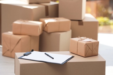 Photo of Clipboard, parcel with tag and blurred stacked boxes on background, indoors