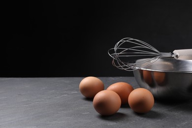 Photo of Metal whisk, raw eggs and bowl on dark grey table. Space for text