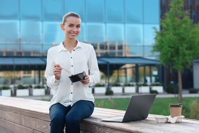 Photo of Smiling businesswoman with lunch box sitting near laptop and paper cup outdoors