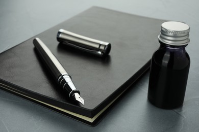 Photo of Stylish fountain pen, bottle of ink and leather notebook on grey textured table, closeup