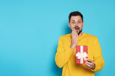 Surprised man with gift box on light blue background, space for text