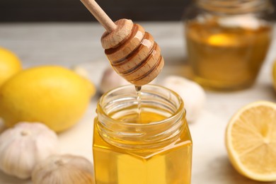 Photo of Honey dripping from dipper into jar, garlic and lemons on table, closeup