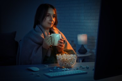 Photo of Beautiful young woman under blanket with popcorn and cup of drink watching film on computer at table indoors at night