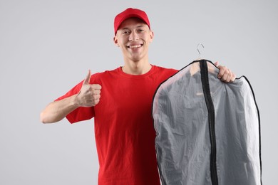 Photo of Dry-cleaning delivery. Happy courier holding garment cover with clothes and showing thumbs up on light grey background