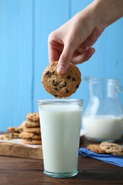 Photo of Woman dipping delicious chocolate chip cookie into glass of milk at wooden table, closeup