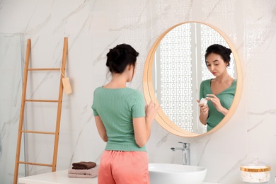 Photo of Young woman applying cream on face near mirror in bathroom
