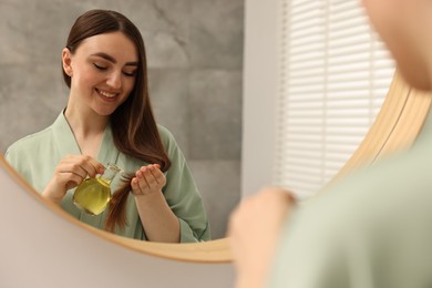 Photo of Young woman applying oil hair mask near mirror at home
