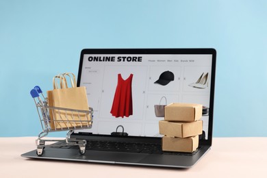 Photo of Online store. Laptop, mini shopping cart and purchases on beige table against light blue background