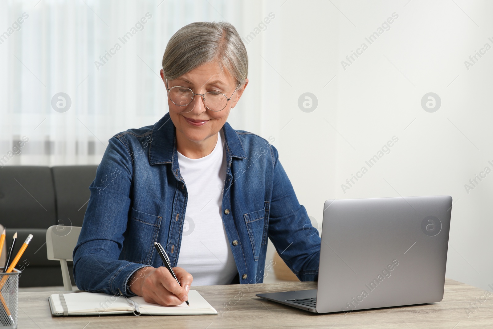 Photo of Beautiful senior woman writing notes in notebook while using laptop at wooden table indoors