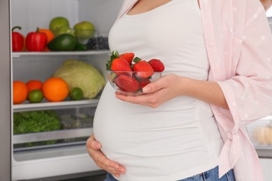Photo of Young pregnant woman with strawberries near fridge at home, closeup. Healthy eating