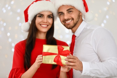 Photo of Happy young couple in Santa hats with gift box against blurred festive lights. Christmas celebration