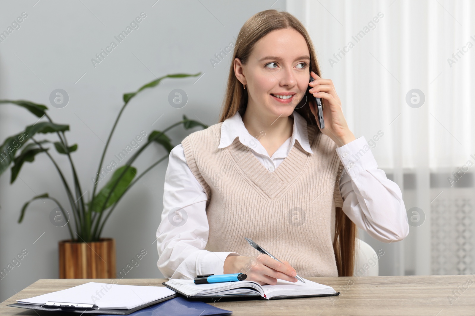 Photo of Woman taking notes while talking on smartphone at wooden table in office