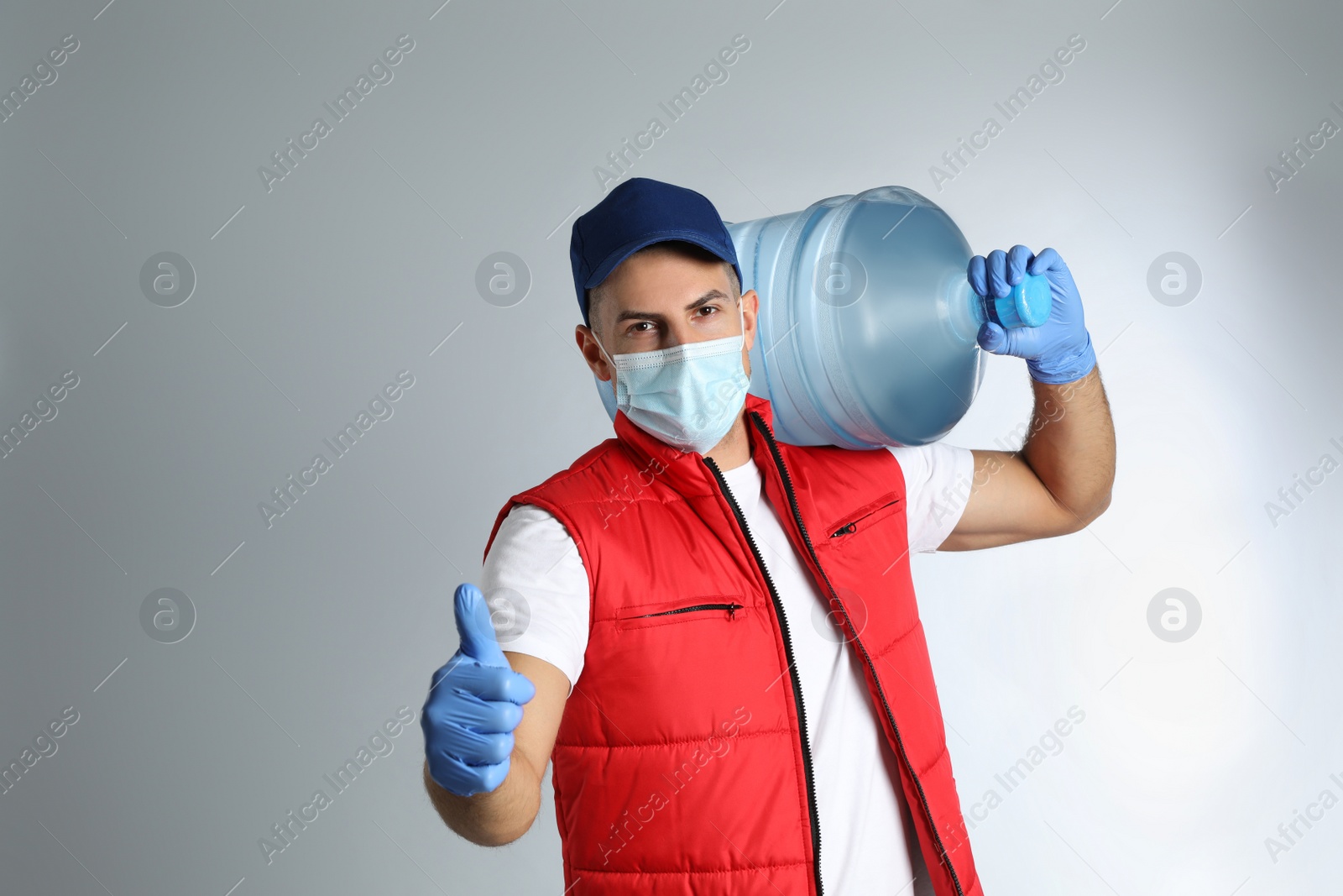 Photo of Courier in face mask with bottle of cooler water on grey background, space for text. Delivery during coronavirus quarantine