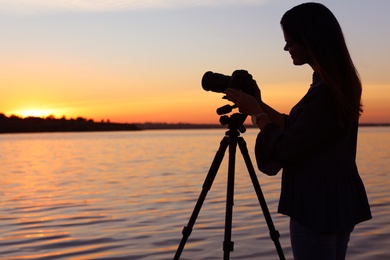 Photo of Young female photographer adjusting professional camera on tripod at sunset