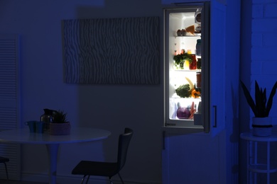 Open refrigerator full of products in stylish kitchen interior at night