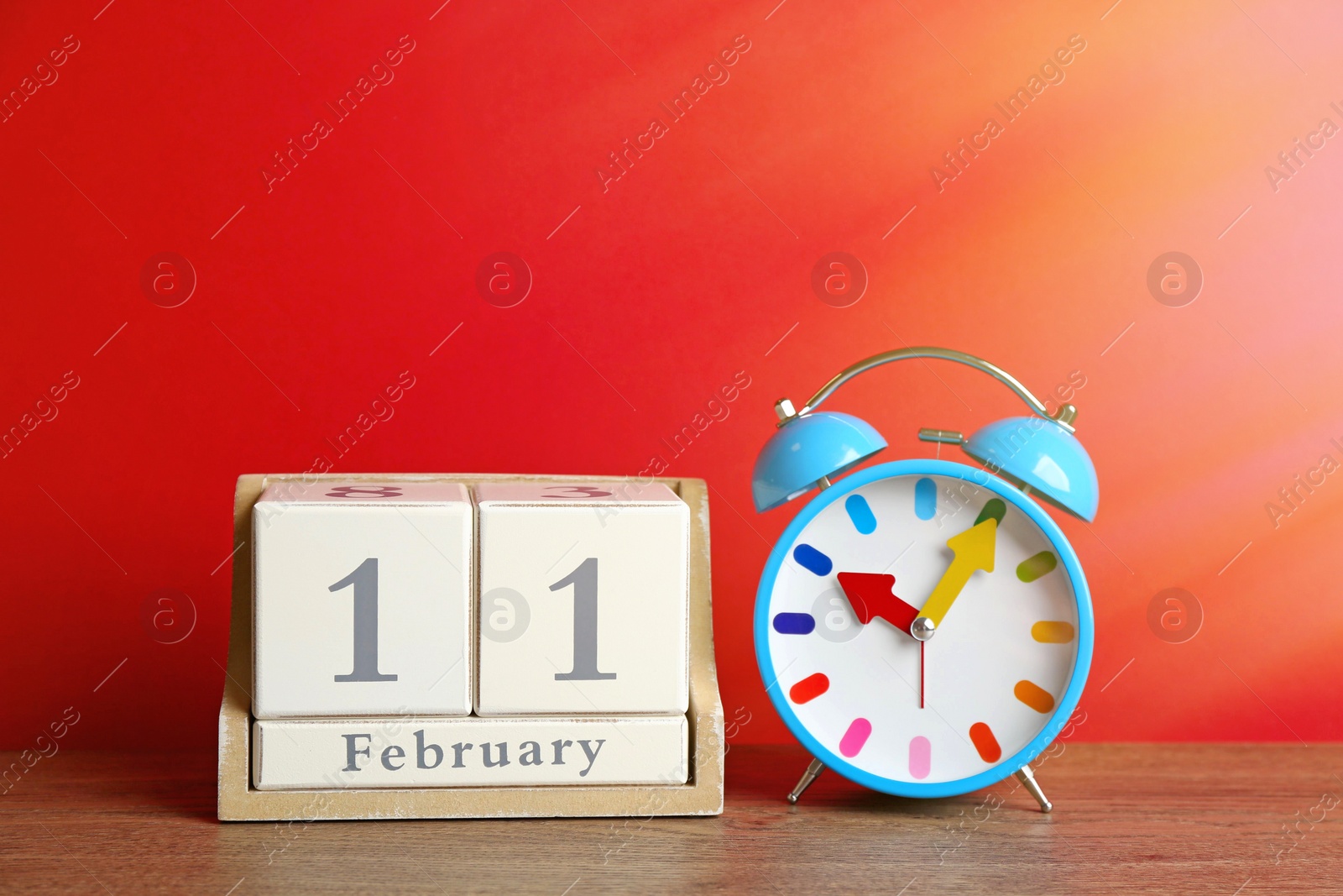 Image of Wooden block calendar and alarm clock on table against red background