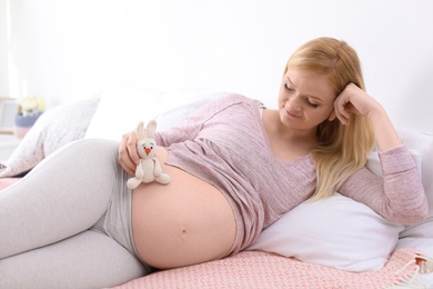 Beautiful pregnant woman holding soft toy near tummy on bed in light room