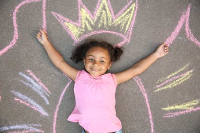 Little African-American child lying near chalk drawing of wings and crown on asphalt, top view