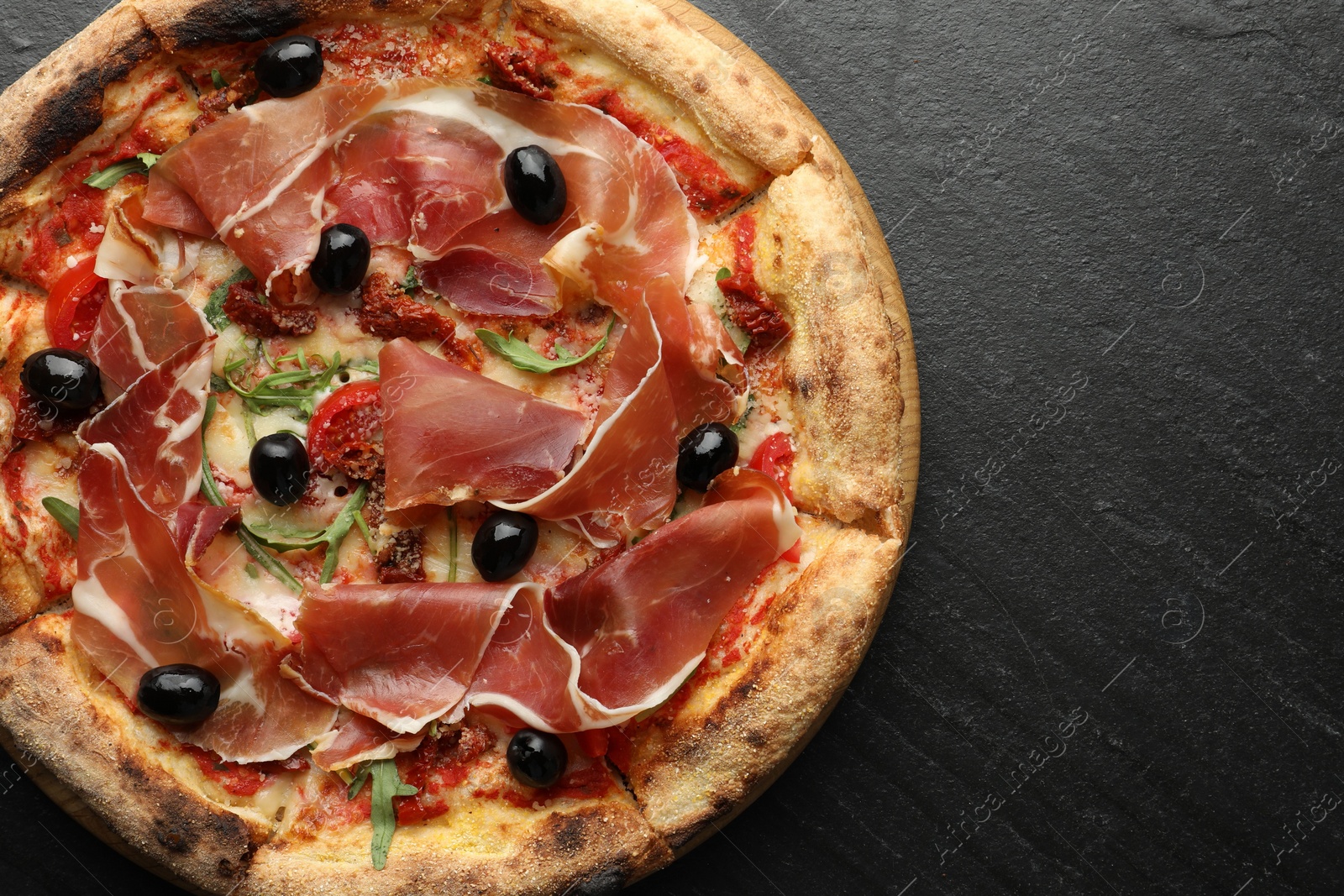 Photo of Tasty pizza with cured ham, olives and sun-dried tomato on black table, top view. Space for text