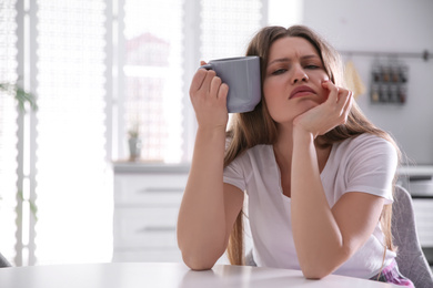 Sleepy young woman with cup of drink at home in morning