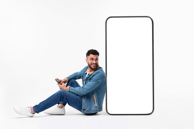 Image of Man with mobile phone sitting near huge device with empty screen on white background. Mockup for design