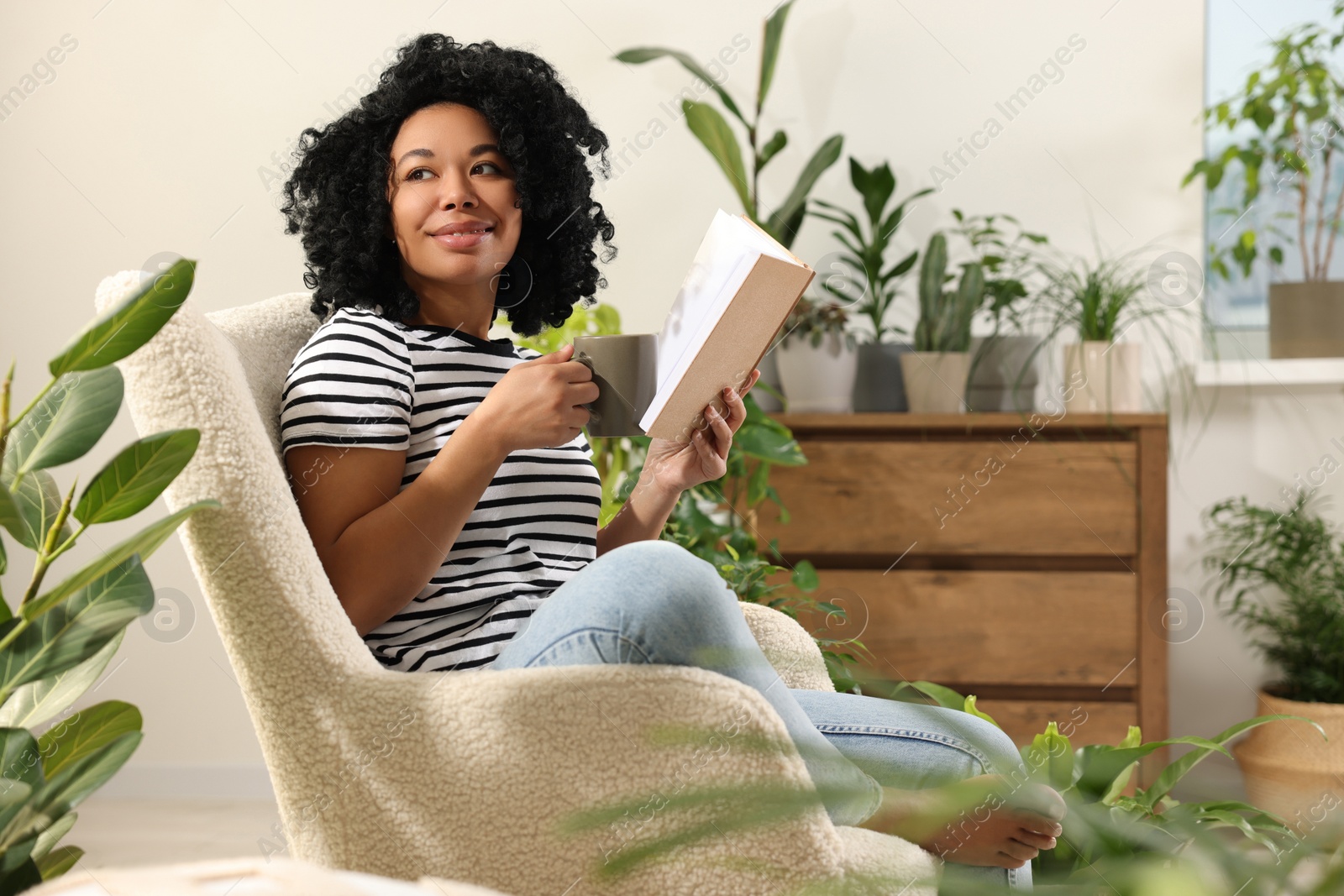 Photo of Relaxing atmosphere. Happy woman with cup of hot drink and book sitting in armchair surrounded by houseplants at home