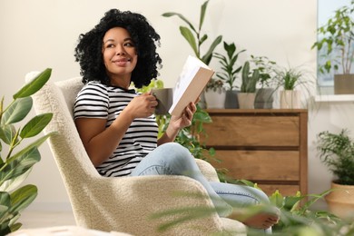 Photo of Relaxing atmosphere. Happy woman with cup of hot drink and book sitting in armchair surrounded by houseplants at home