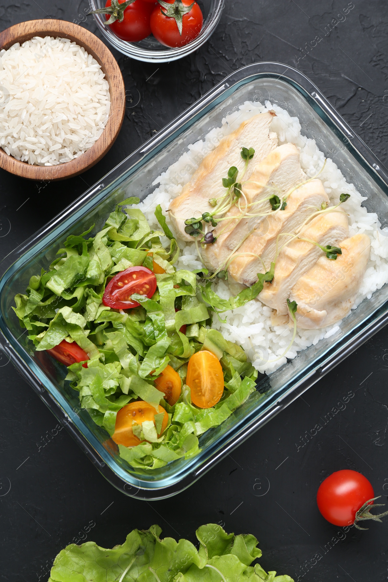 Photo of Healthy meal. Fresh salad, chicken and rice in glass container near other products on black table, flat lay