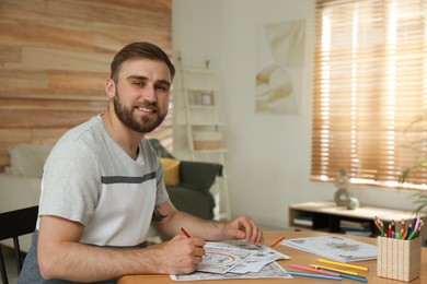 Young man coloring antistress picture at table indoors