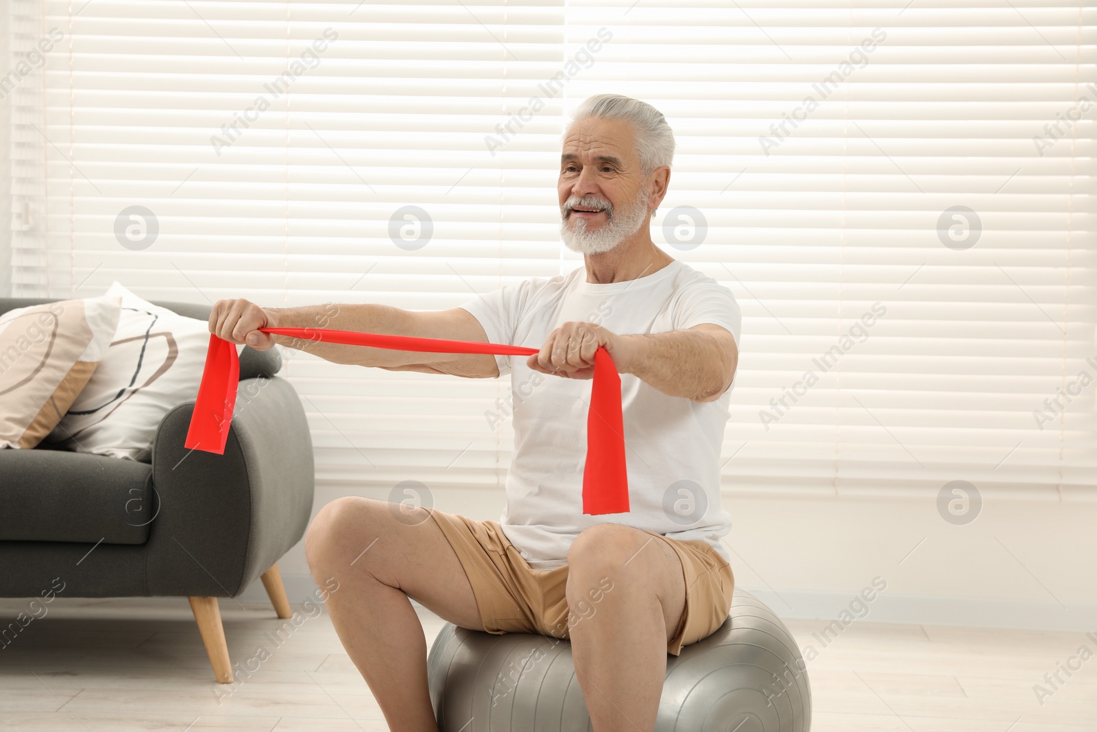 Photo of Senior man doing exercise with elastic resistance band on fitness ball at home