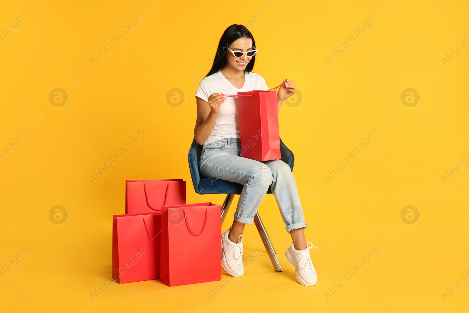 Photo of Beautiful young woman with paper shopping bags in armchair on yellow background