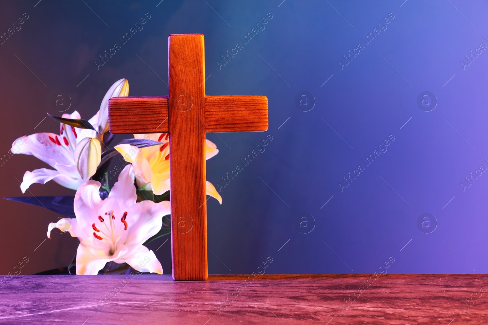 Photo of Wooden cross and beautiful lily flowers on table in color lights, space for text. Religion of Christianity
