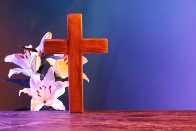 Wooden cross and beautiful lily flowers on table in color lights, space for text. Religion of Christianity