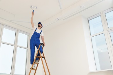 Photo of Worker in uniform painting ceiling with roller on stepladder indoors, low angle view