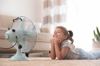 Photo of Little girl enjoying air flow from fan on floor in living room. Summer heat