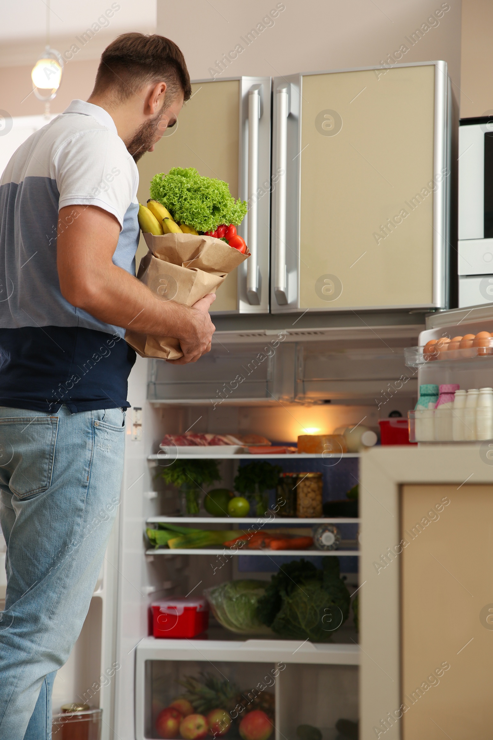 Photo of Young man with fresh food in paper bag near refrigerator at home