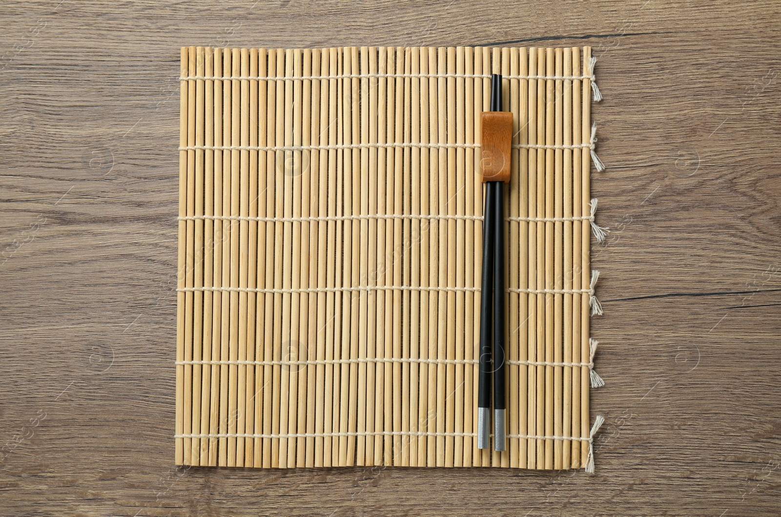 Photo of Bamboo mat with pair of black chopsticks and rest on wooden table, top view