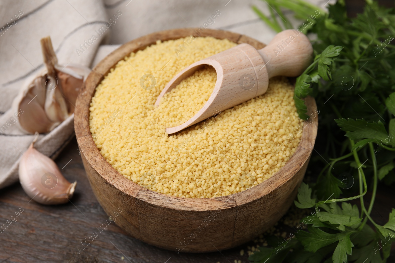 Photo of Raw couscous in bowl, scoop, parsley and garlic on table, closeup