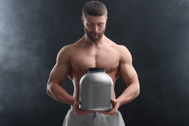 Young man with muscular body holding jar of protein powder on dark grey background