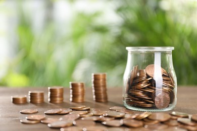 Glass jar with metal coins on wooden table against blurred green background. Space for text