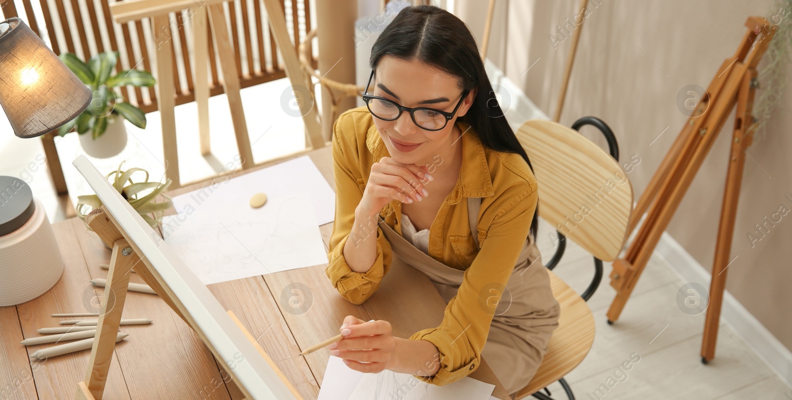 Image of Young woman drawing on easel with pencil at table indoors, above view. Banner design