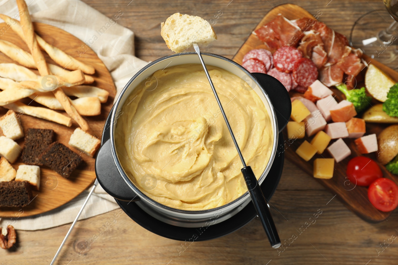 Photo of Fork with piece of bread, melted cheese in fondue pot and snacks on wooden table, flat lay