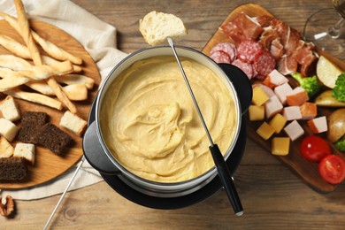 Fork with piece of bread, melted cheese in fondue pot and snacks on wooden table, flat lay