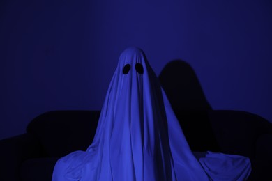 Photo of Creepy ghost. Woman covered with sheet sitting on sofa in blue light