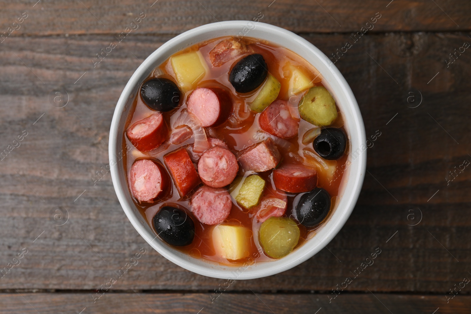 Photo of Meat solyanka soup with thin dry smoked sausages in bowl on wooden table, top view