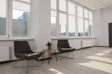 Modern armchairs and table near large windows with white roller blinds indoors