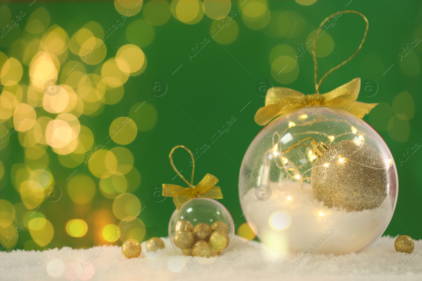 Image of Beautiful transparent Christmas ornaments with golden bows on snow against green background, bokeh effect. Space for text