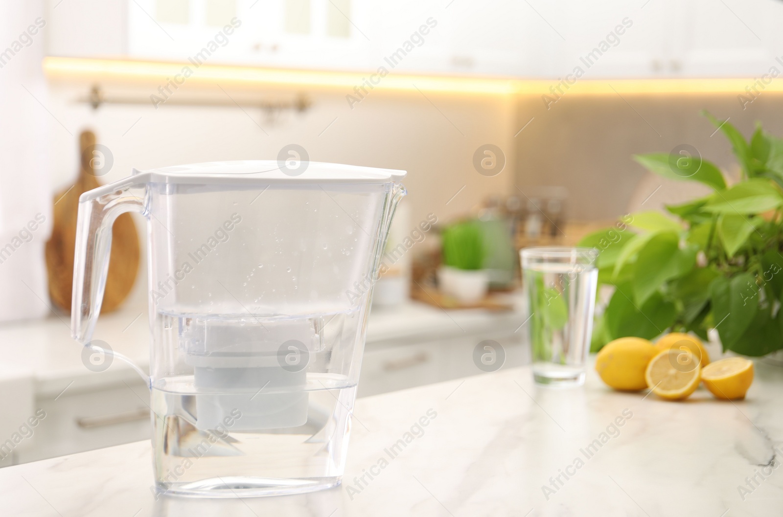 Photo of Water filter jug, glass and lemons on white marble table in kitchen